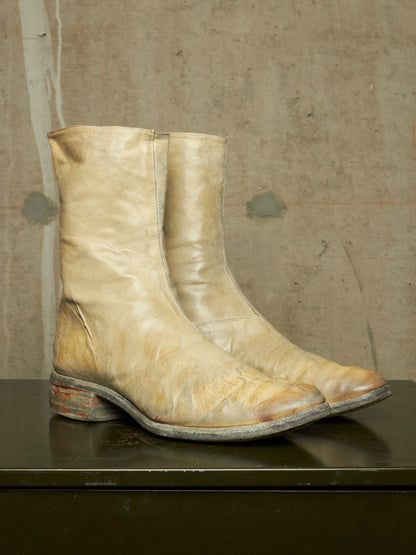A pair of distressed light beige leather ankle boots with a western design, resting on a dark surface against a textured concrete wall.