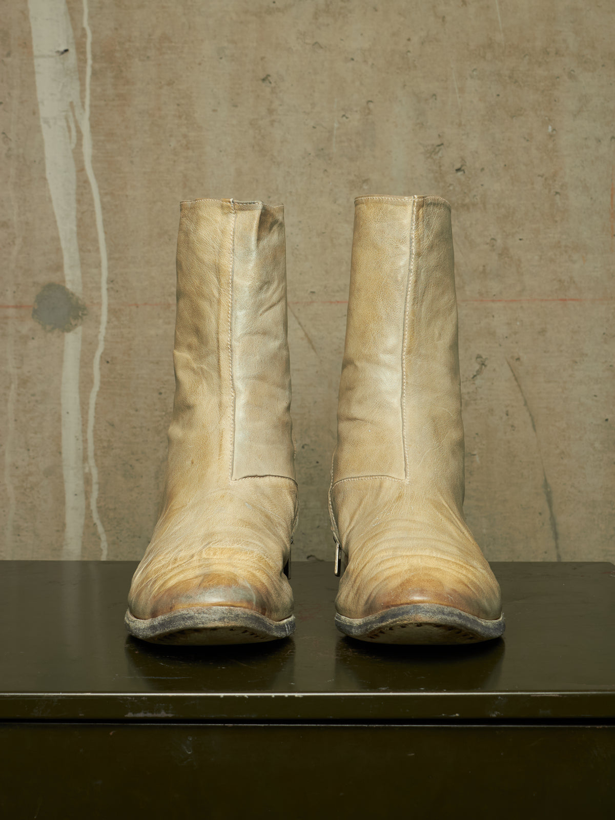 A pair of worn beige leather ankle boots with a vertical seam down the front, resting on a dark surface against a textured concrete wall.