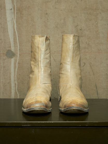 A pair of worn beige leather ankle boots with a vertical seam down the front, resting on a dark surface against a textured concrete wall.