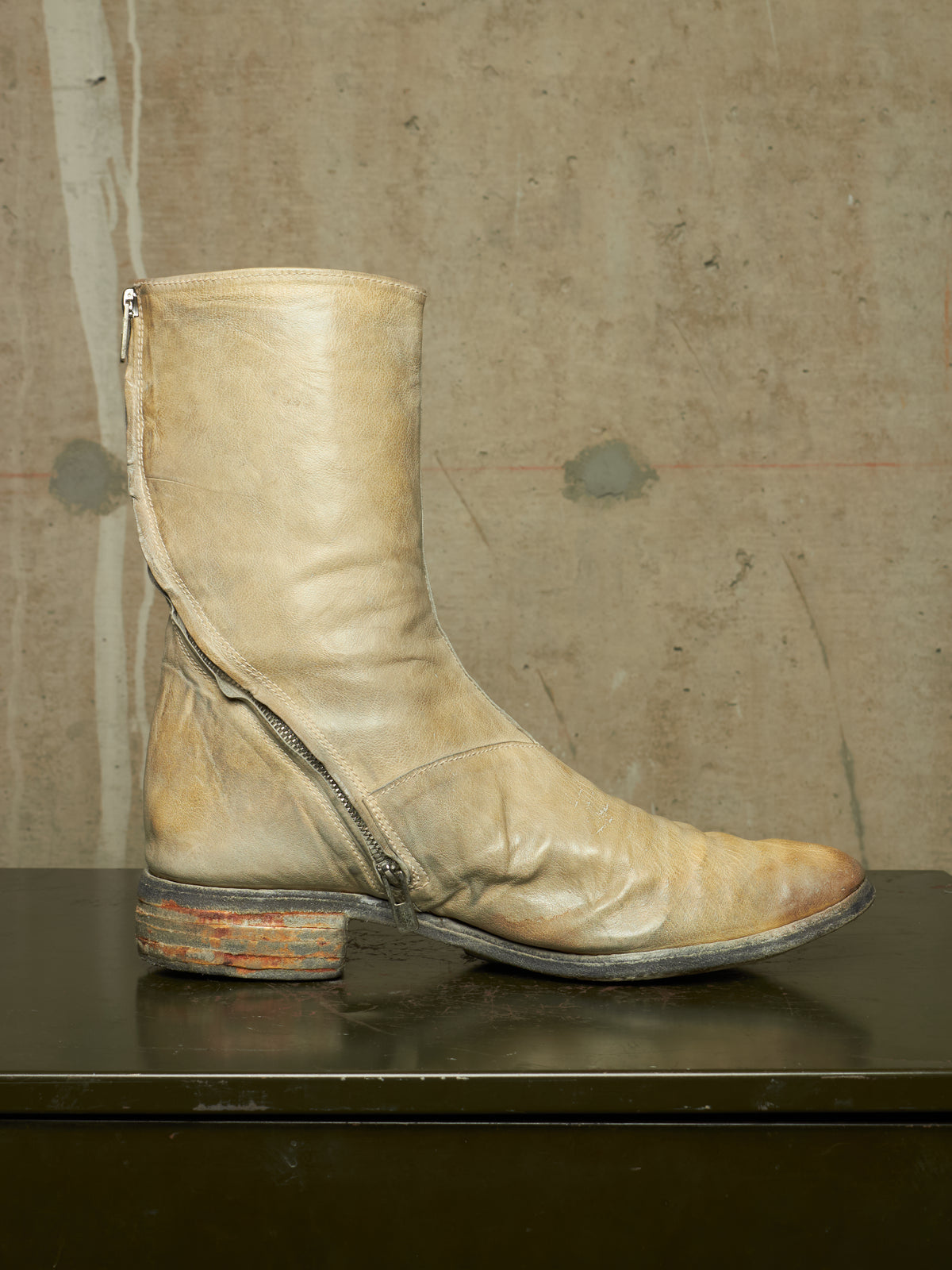 A single light beige leather ankle boot with a side zipper, displayed in profile on a dark reflective surface against a textured concrete wall.