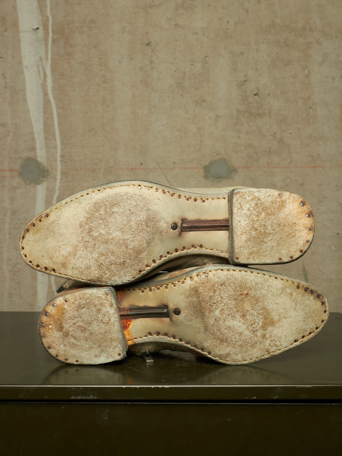 A pair of worn leather shoes shown from the sole side, resting on a dark surface against a textured concrete wall.