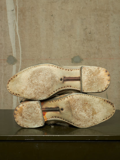 A pair of worn leather shoes shown from the sole side, resting on a dark surface against a textured concrete wall.