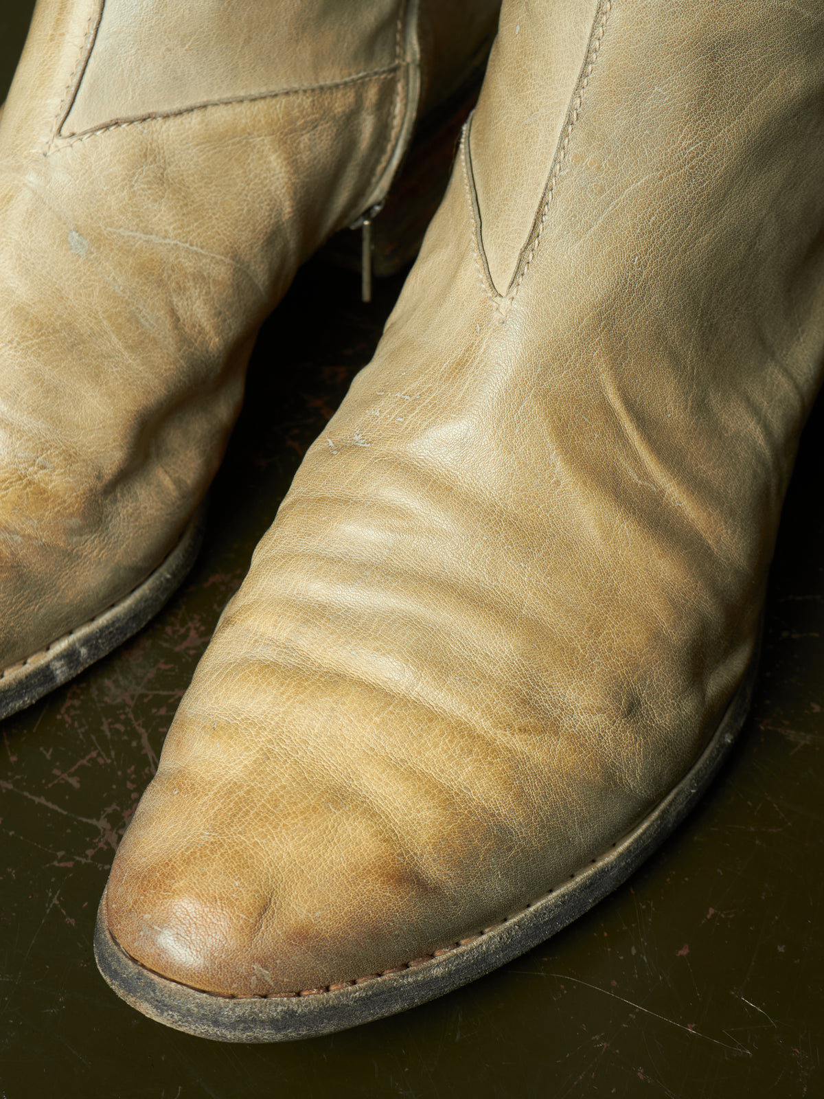 A close-up of a pair of worn beige leather cowboy boots with visible scuffs, creases, and dirt on a dark reflective surface.