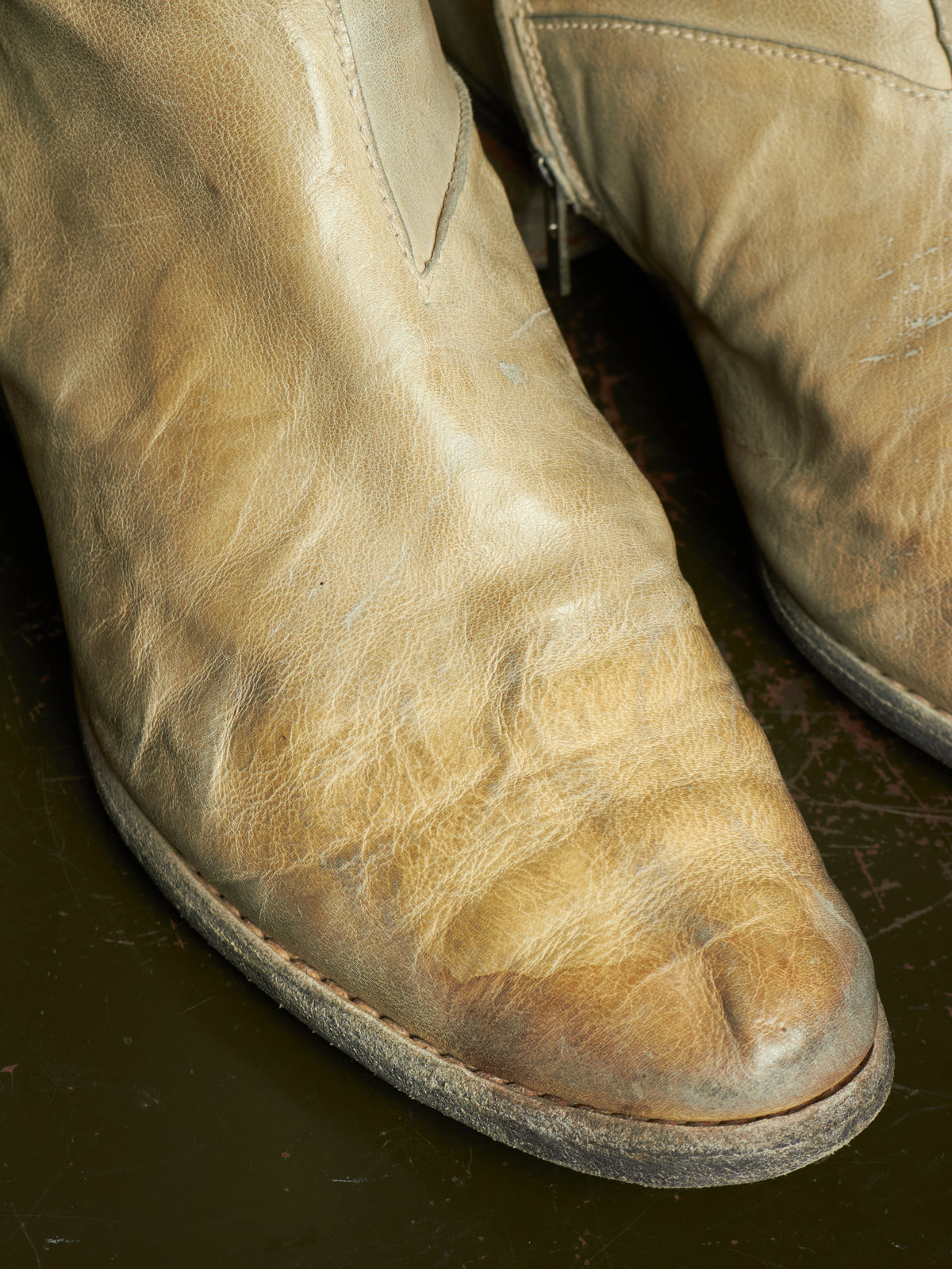 A close-up of a heavily worn, distressed tan leather cowboy boot bootie, showing significant scuffing and creasing on the toe and shaft, with another boot partially visible in the background.