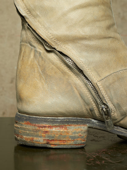 Close-up of a distressed beige leather boot with a side zipper and a weathered stacked-wood-look heel showing rust and gray tones.