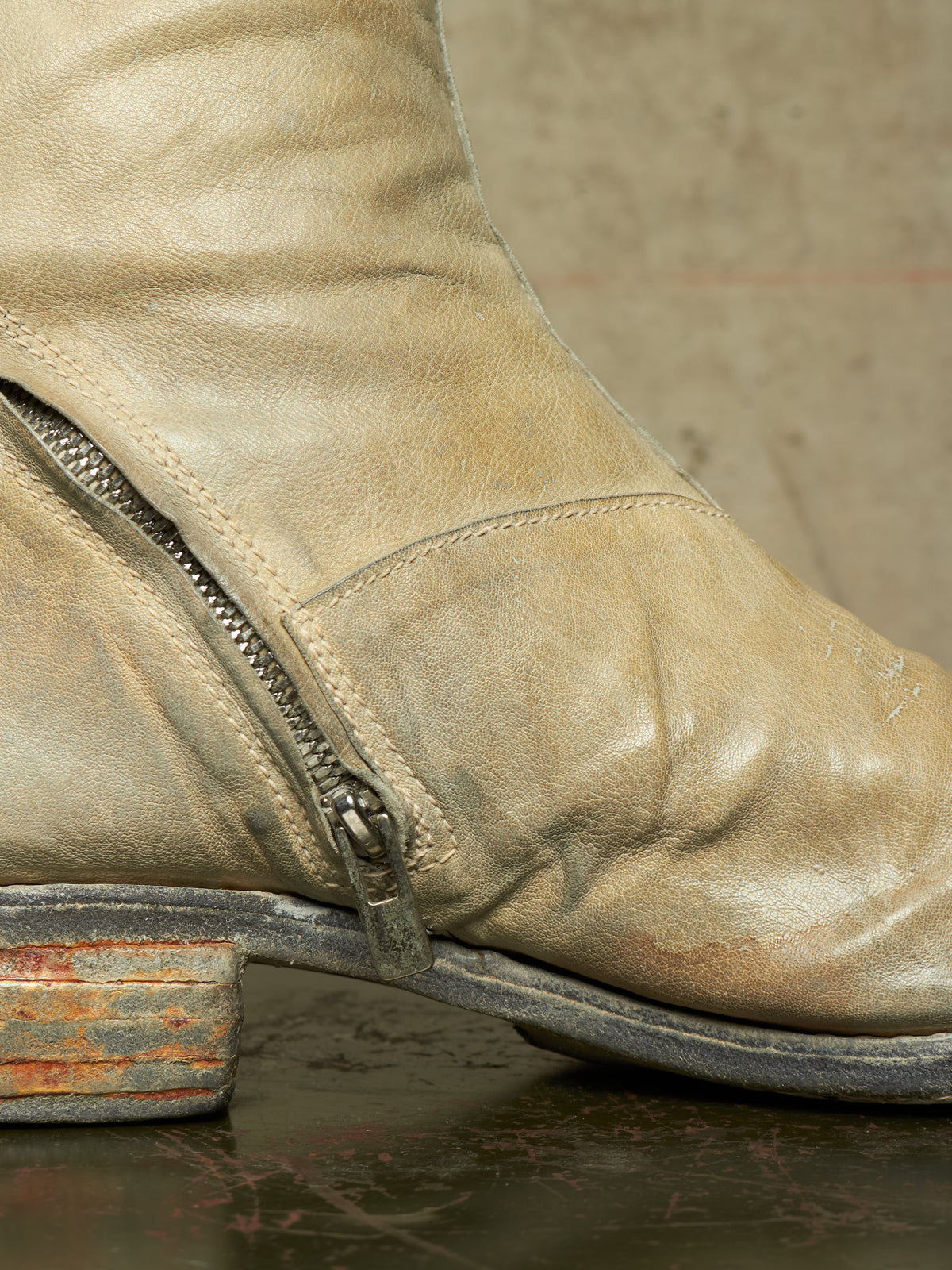 Close-up of a distressed beige leather boot with a side zipper, a rustic distressed block heel, and a dark sole, resting on a dark surface against a textured neutral background.