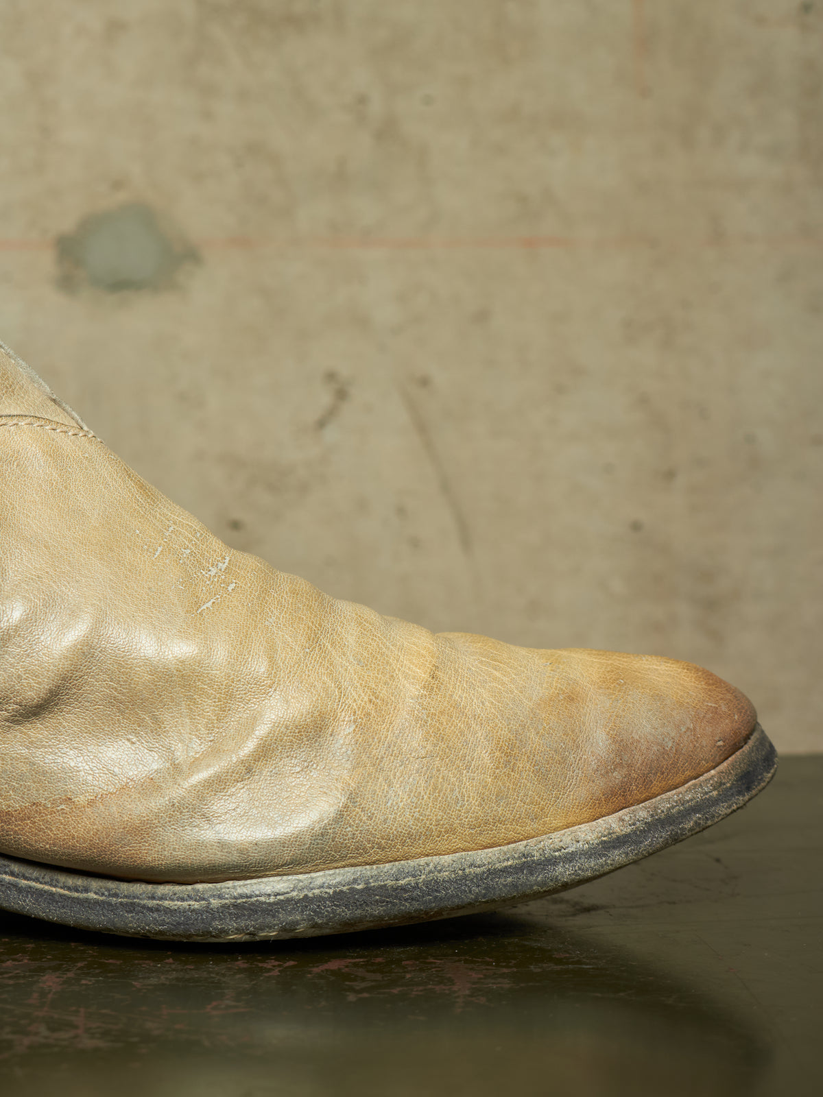 A close-up of a worn, light tan leather boot with a dark sole, resting on a dark reflective surface against a textured beige wall.