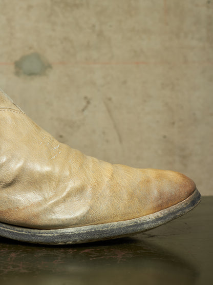 A close-up of a worn, light tan leather boot with a dark sole, resting on a dark reflective surface against a textured beige wall.