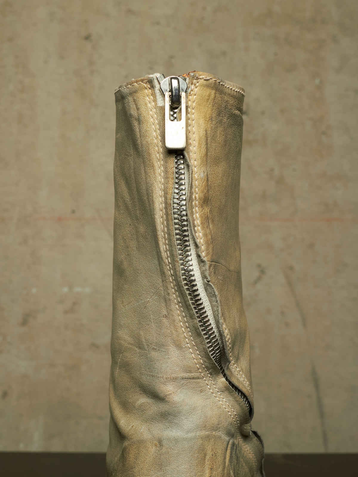 A close-up of a distressed beige leather boot featuring a prominent silver metal zipper running down the center, set against a textured concrete background.