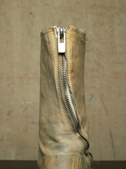 A close-up of a distressed beige leather boot featuring a prominent silver metal zipper running down the center, set against a textured concrete background.