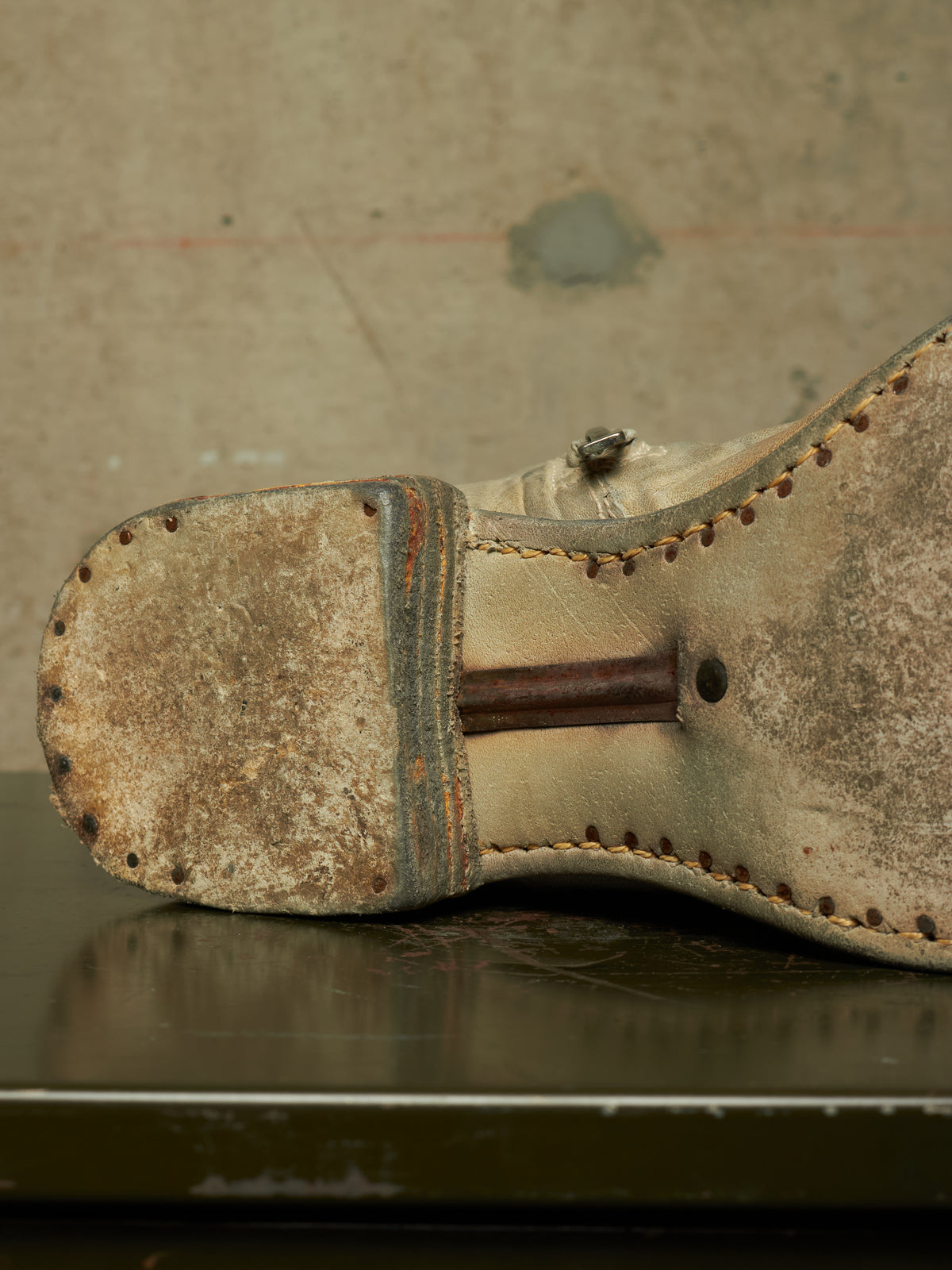 A close-up of the sole of a worn, vintage beige leather shoe or boot, showing heavy scuffing, a metal heel pin, and decorative stitching, resting on a dark reflective surface against a textured concrete background.