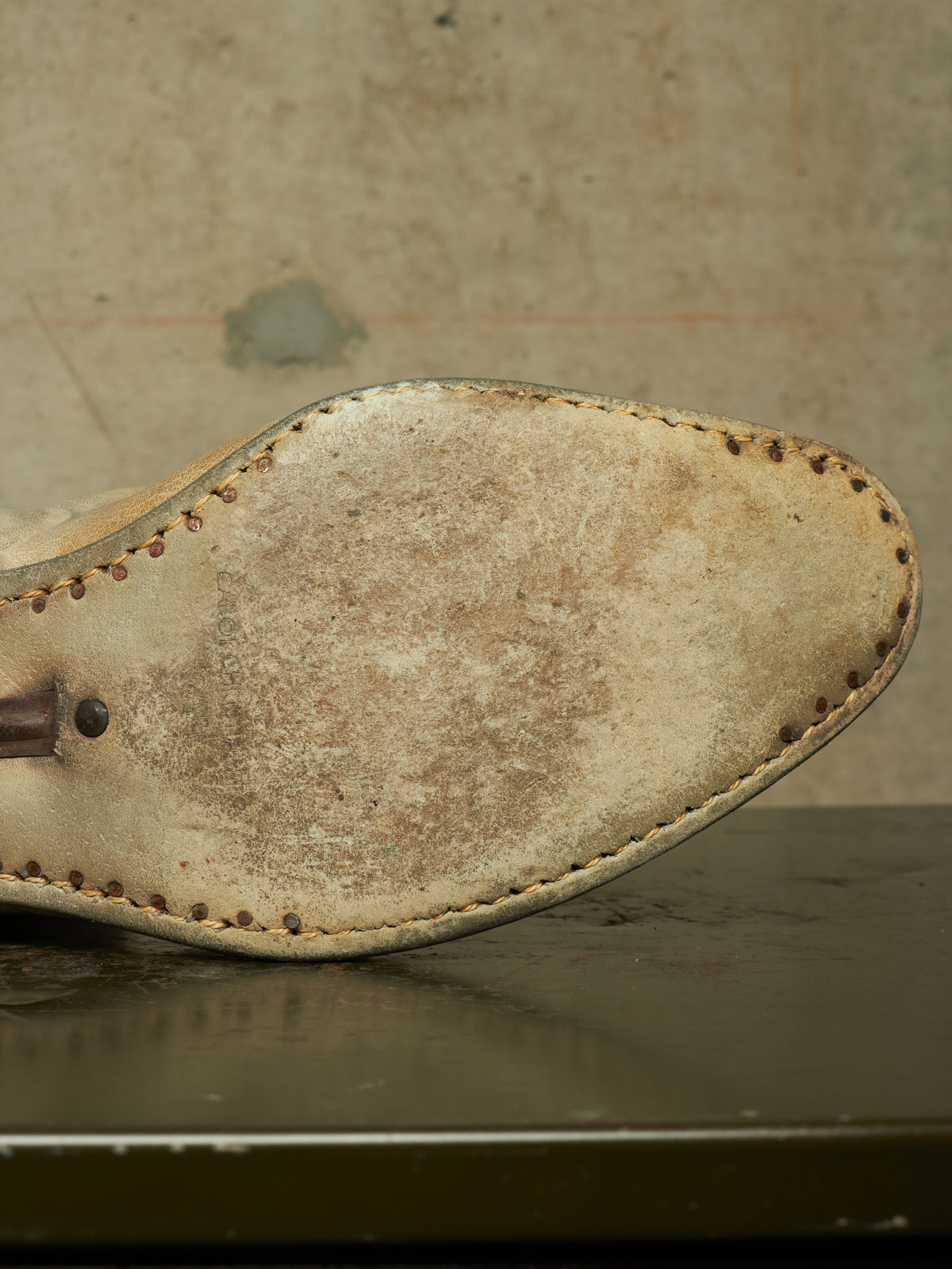 Close-up of the worn underside of a light tan leather shoe or boot, showing the sole, stitching, and a small metal rivet against a textured beige background.