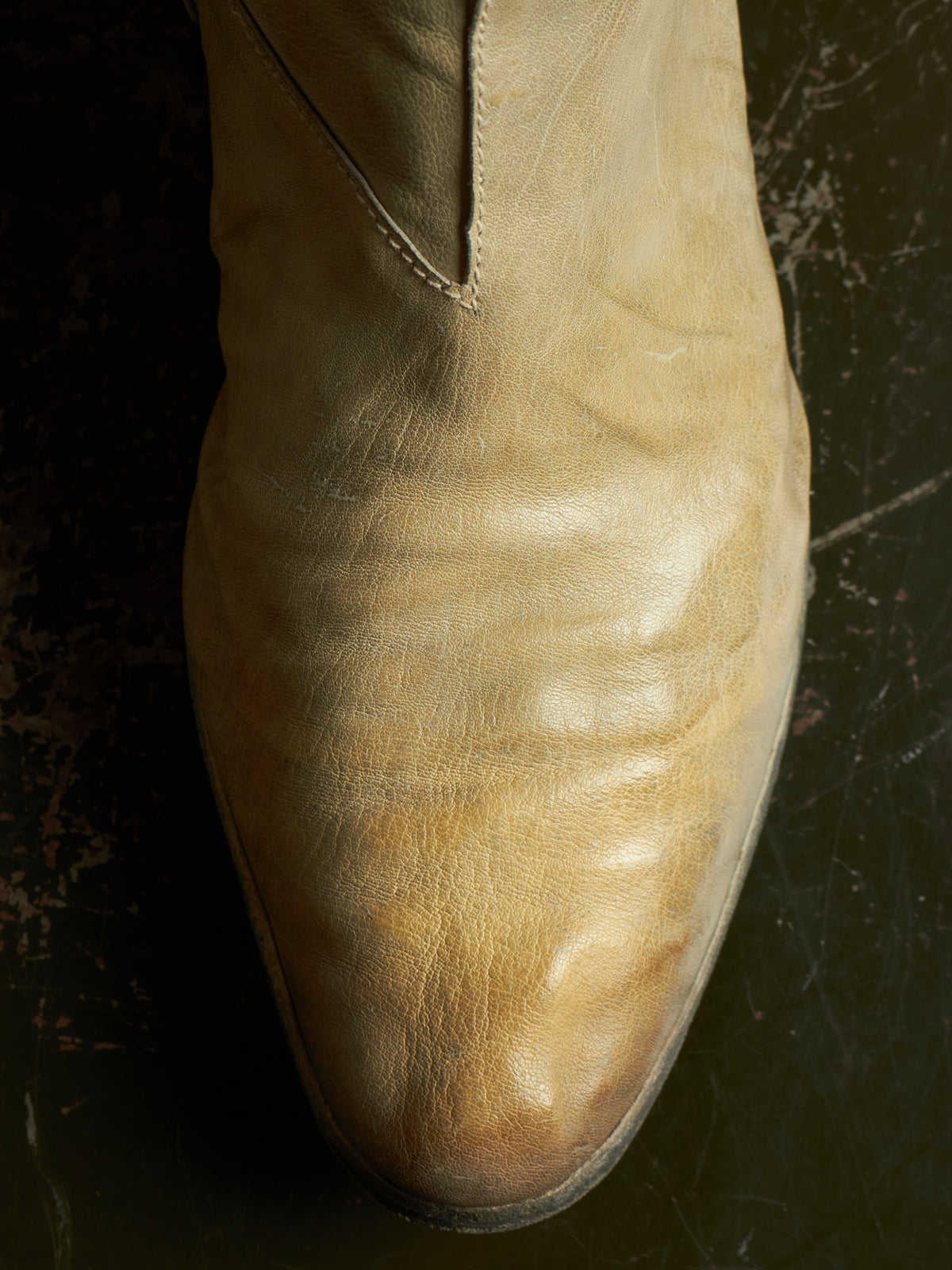 A close-up of a light tan leather boot with a pointed toe, showing the texture and creasing of the leather against a dark, textured background.
