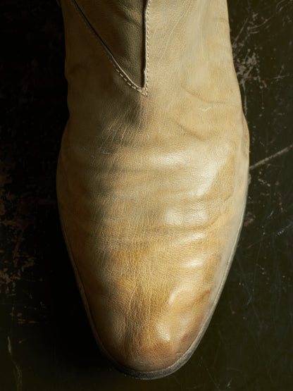 A close-up of a light tan leather boot with a pointed toe, showing the texture and creasing of the leather against a dark, textured background.