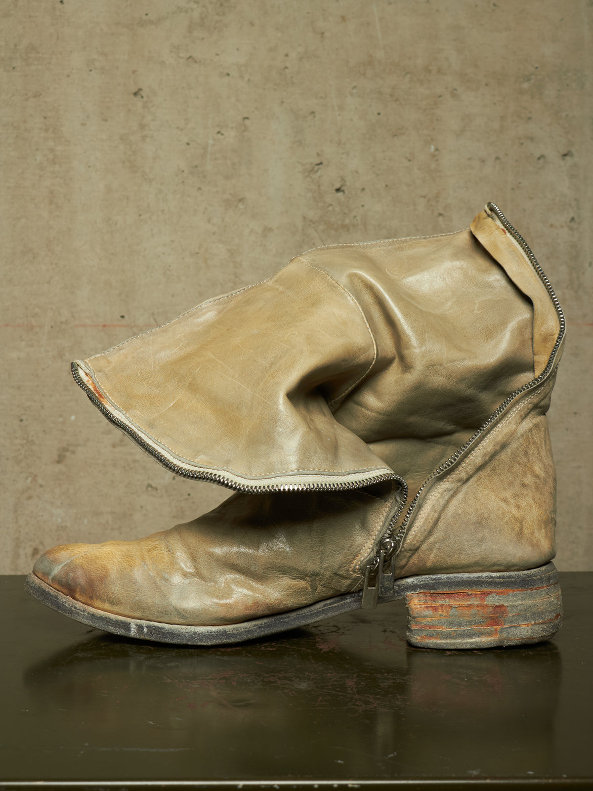 A single distressed leather ankle boot with a side zipper, shown in a side profile against a textured concrete wall background.