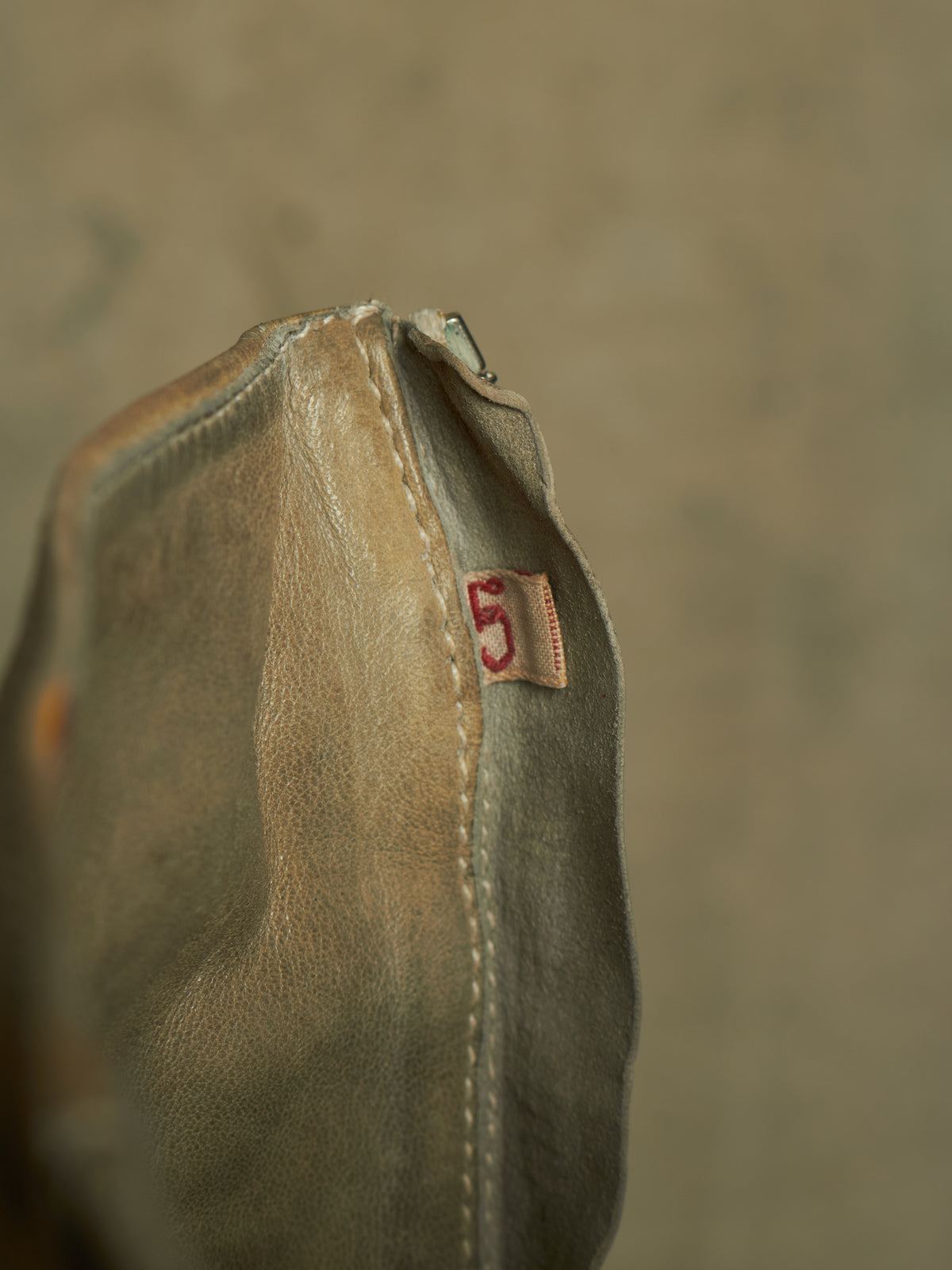 A close-up of a distressed leather pouch or bag corner showing a small tan leather tag with a red embroidered number 5, set against a textured beige background.