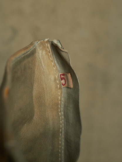 A close-up of a distressed leather pouch or bag corner showing a small tan leather tag with a red embroidered number 5, set against a textured beige background.