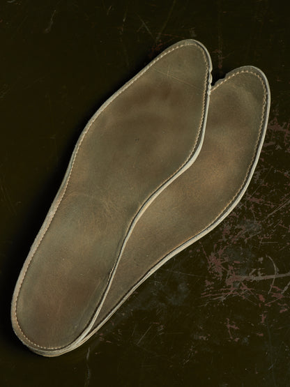 A pair of unfinished leather shoe insoles with visible stitching, displayed on a dark textured background.