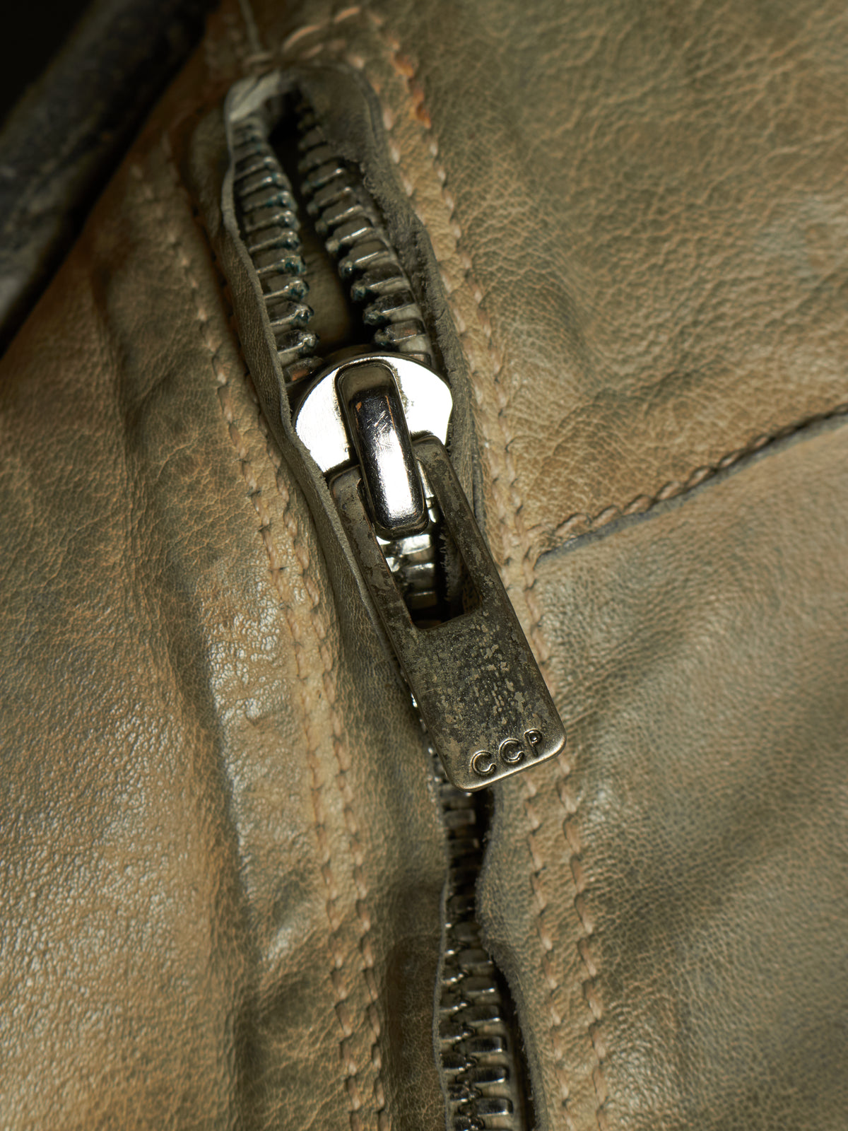 Close-up of a brown leather jacket with a silver metal zipper pulled open, showing the texture of the leather and the zipper pull.