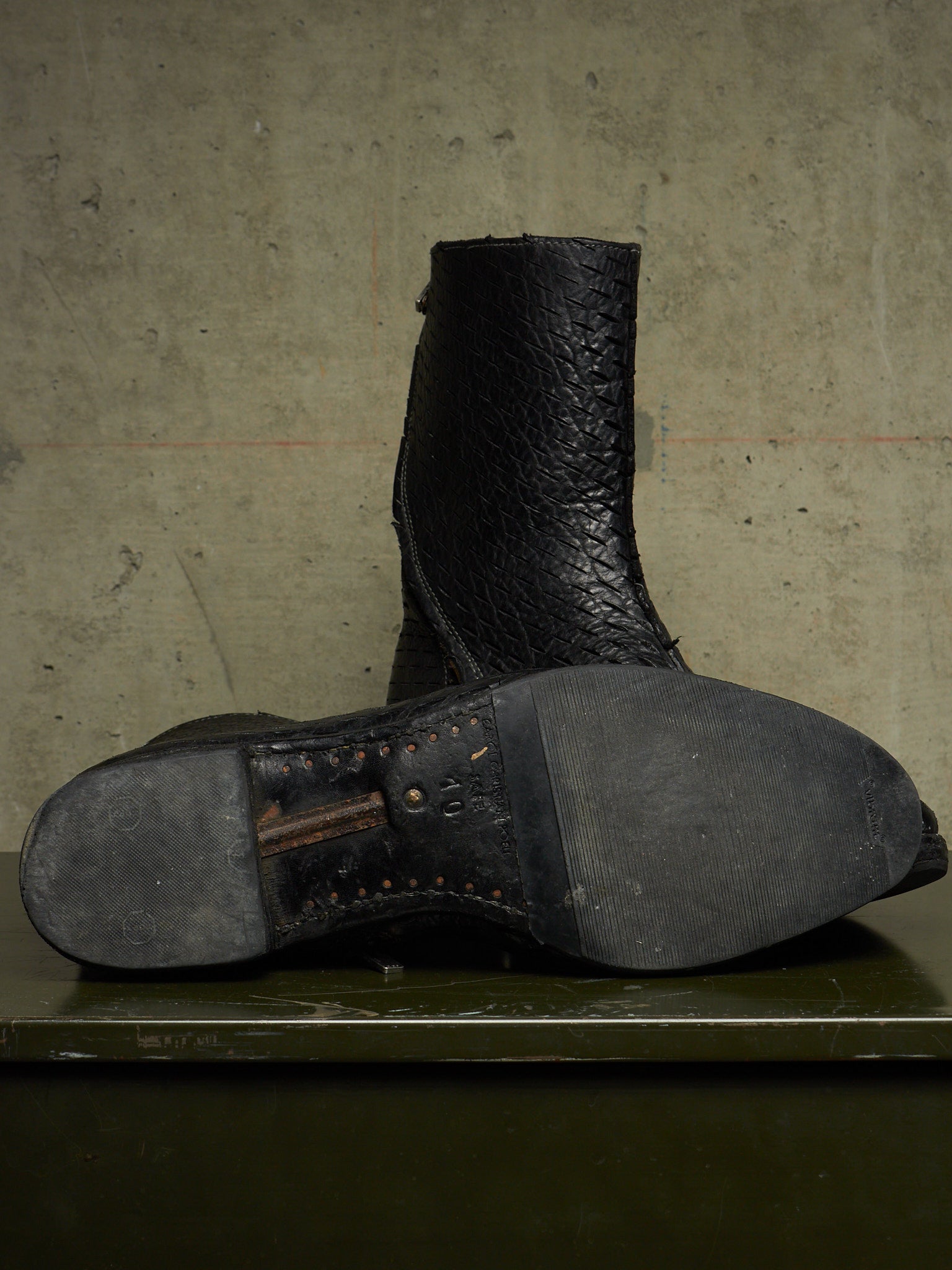 A single black leather boot shown from the sole and heel side, resting on a dark surface against a textured concrete wall.