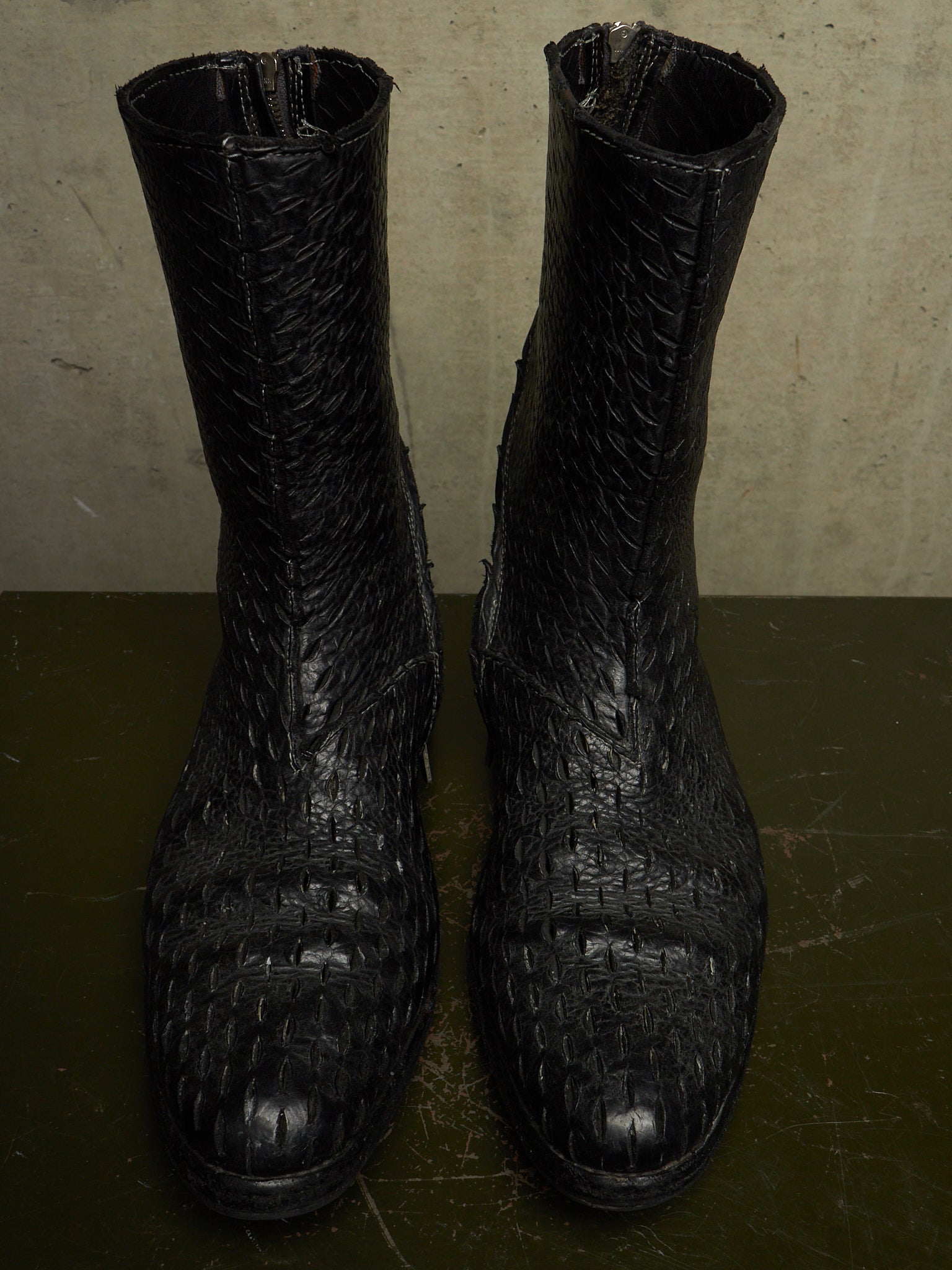 A pair of black crocodile-embossed leather ankle boots with a textured, woven pattern, shown against a neutral concrete background.