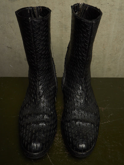 A pair of black crocodile-embossed leather ankle boots with a textured, woven pattern, shown against a neutral concrete background.
