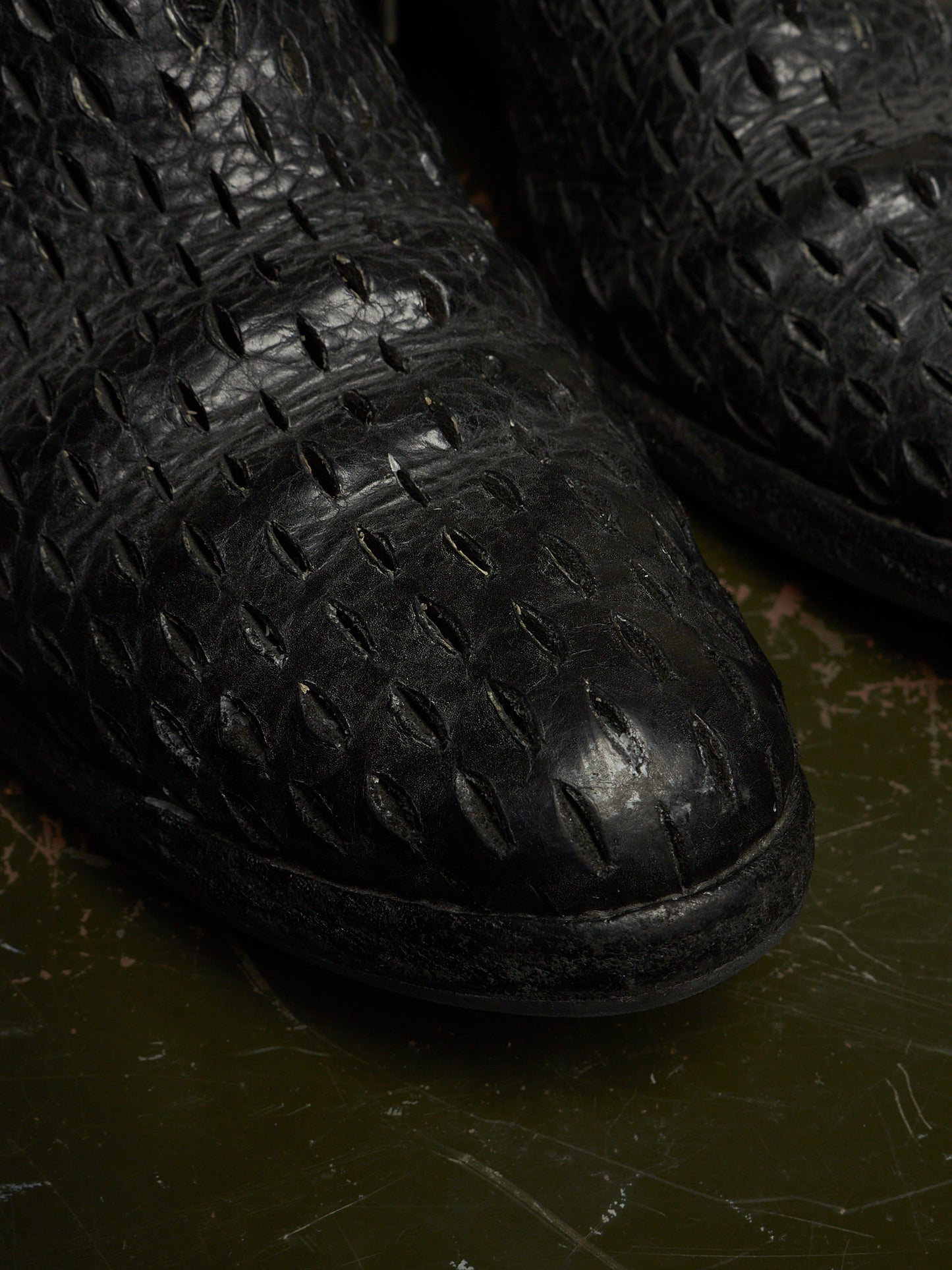Close-up of a black crocodile-embossed leather boot toe showing detailed texture and a dark green textured surface underneath.