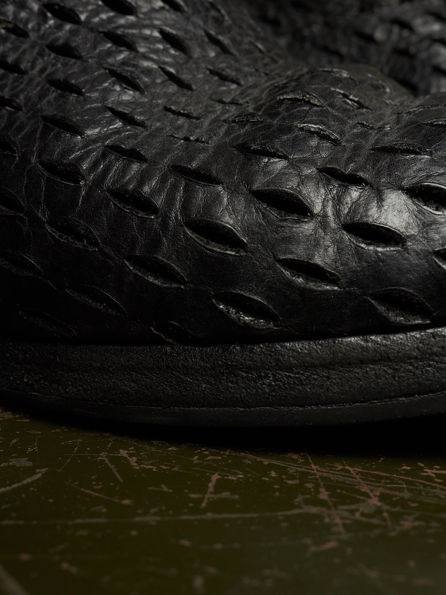Close-up of a black textured leather shoe with a unique embossed pattern, resting on a dark olive green surface.