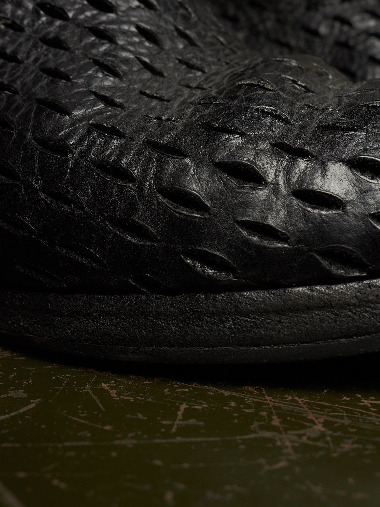 Close-up of a black textured leather shoe with a unique embossed pattern, resting on a dark olive green surface.