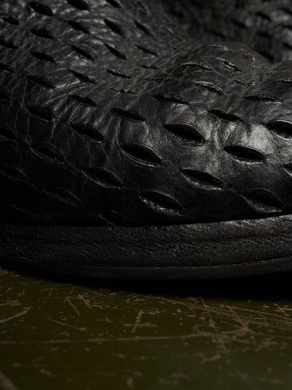 Close-up of a black textured leather shoe with a unique embossed pattern, resting on a dark olive green surface.