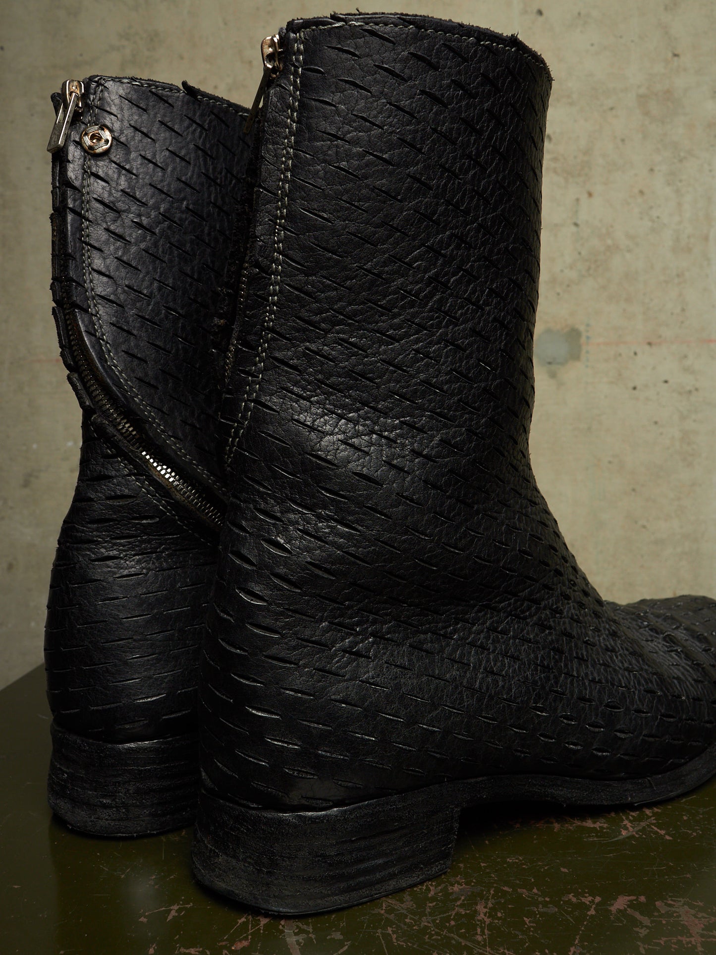 A pair of black leather ankle boots with a distinctive embossed basketweave texture, featuring side zippers, shown on a dark green surface against a textured beige wall.