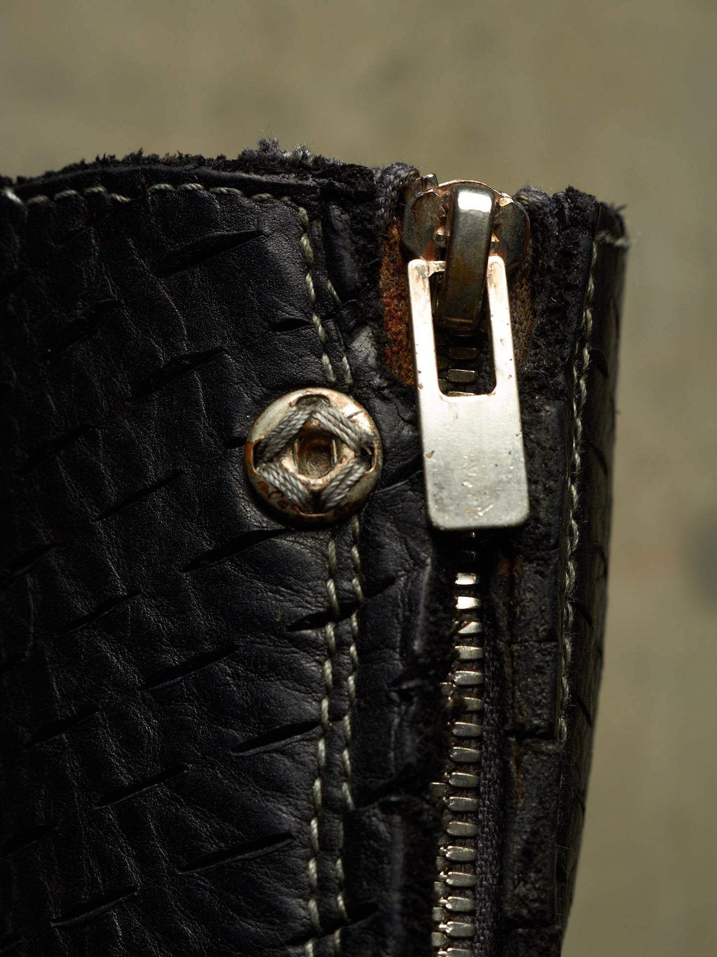 Close-up of a black woven leather zipper boot with a silver metal zipper pull and a decorative metal rivet.