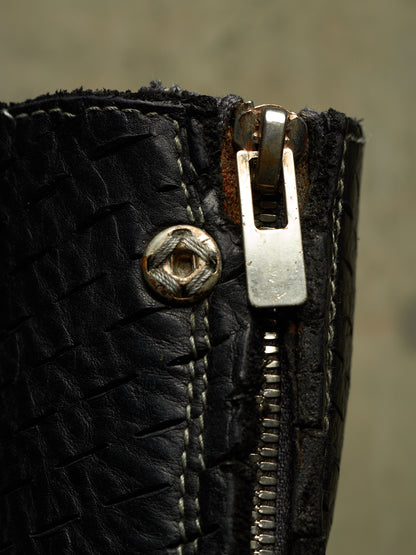Close-up of a black woven leather zipper boot with a silver metal zipper pull and a decorative metal rivet.