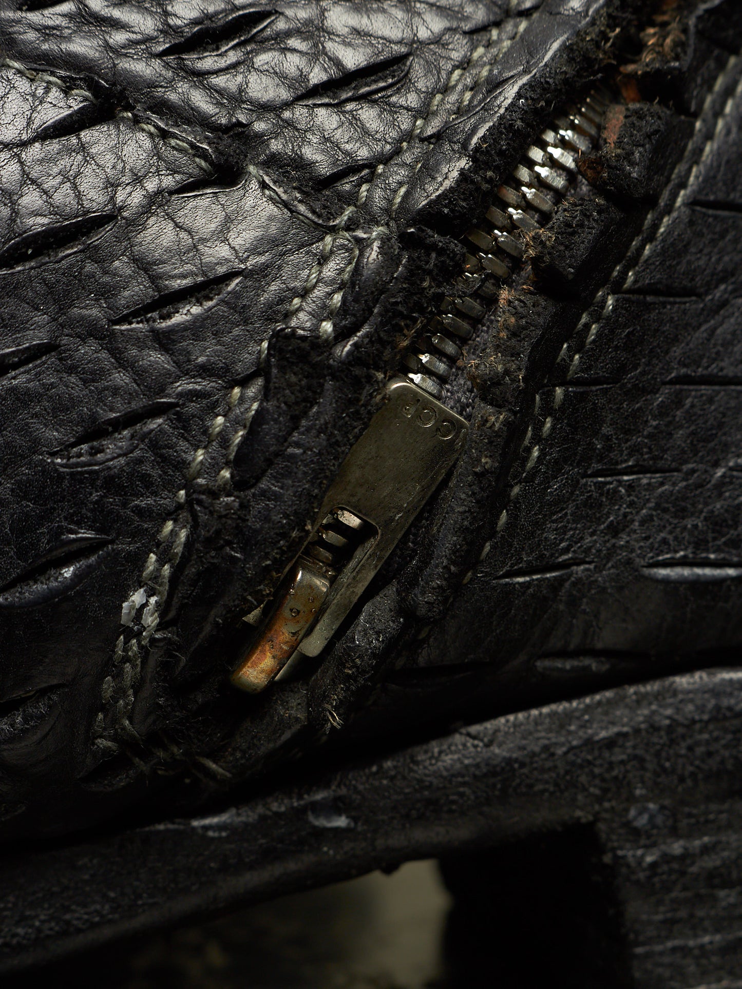 Extreme close-up of a black textured leather zipper, showing the black metal zipper pull with rust and dirt, white stitching, and the textured black leather surface.