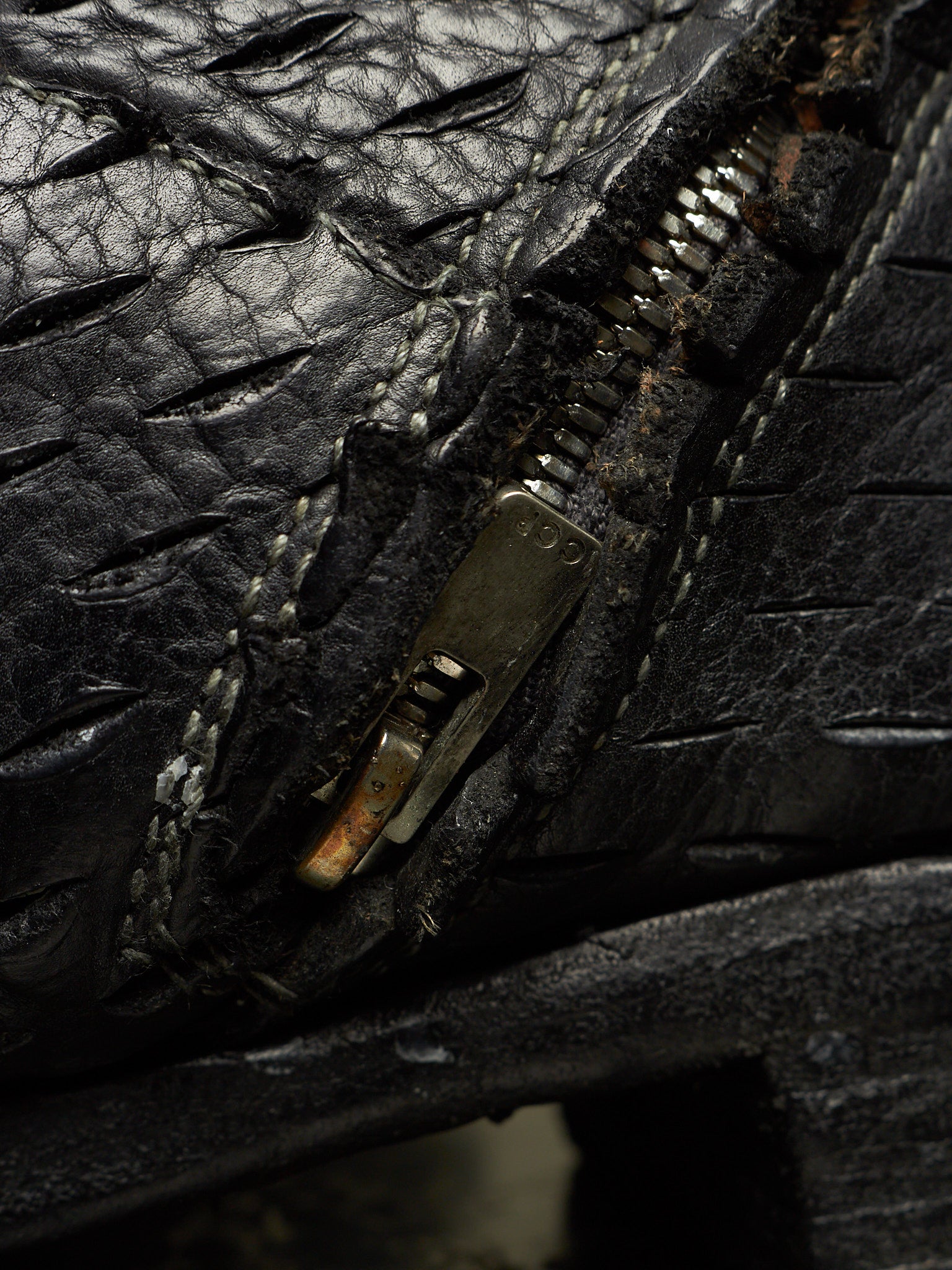 Extreme close-up of a black textured leather zipper, showing the black metal zipper pull with rust and dirt, white stitching, and the textured black leather surface.