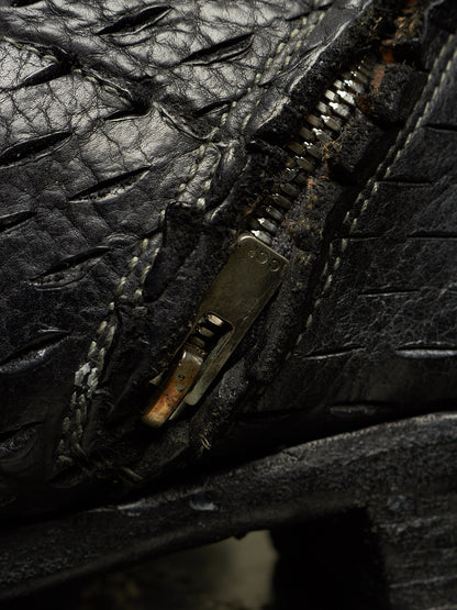 Extreme close-up of a black textured leather zipper, showing the black metal zipper pull with rust and dirt, white stitching, and the textured black leather surface.