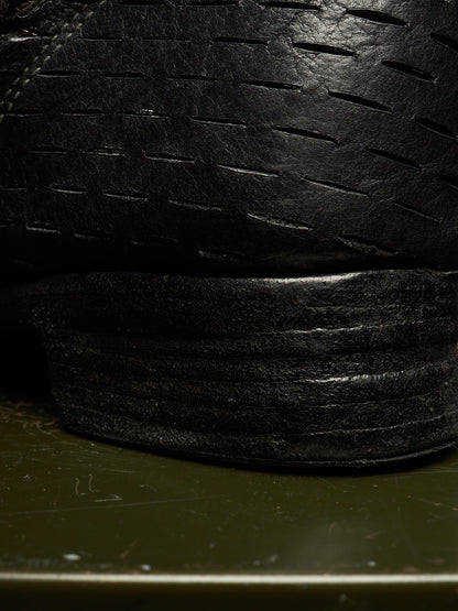 Close-up of a black leather shoe sole and textured upper with stitched detailing, photographed against a dark olive background.