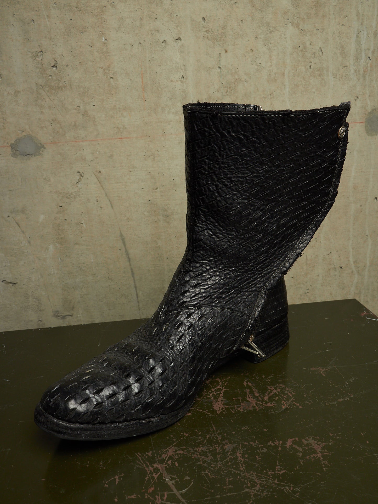 A single black leather ankle boot with a textured, crocodile-embossed toe and shaft, resting on a dark surface against a concrete wall.
