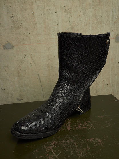 A single black leather ankle boot with a textured, crocodile-embossed toe and shaft, resting on a dark surface against a concrete wall.