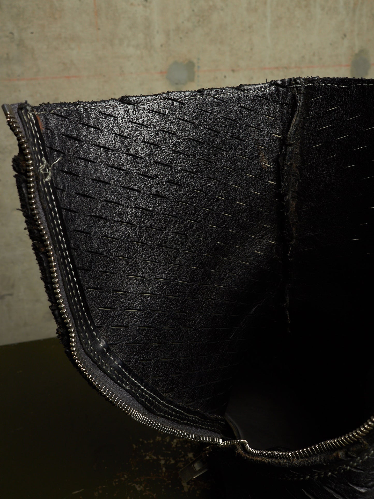 Close-up of a black woven-texture leather accessory, likely a bag or pouch, showing the zipper and interior lining against a concrete wall background.