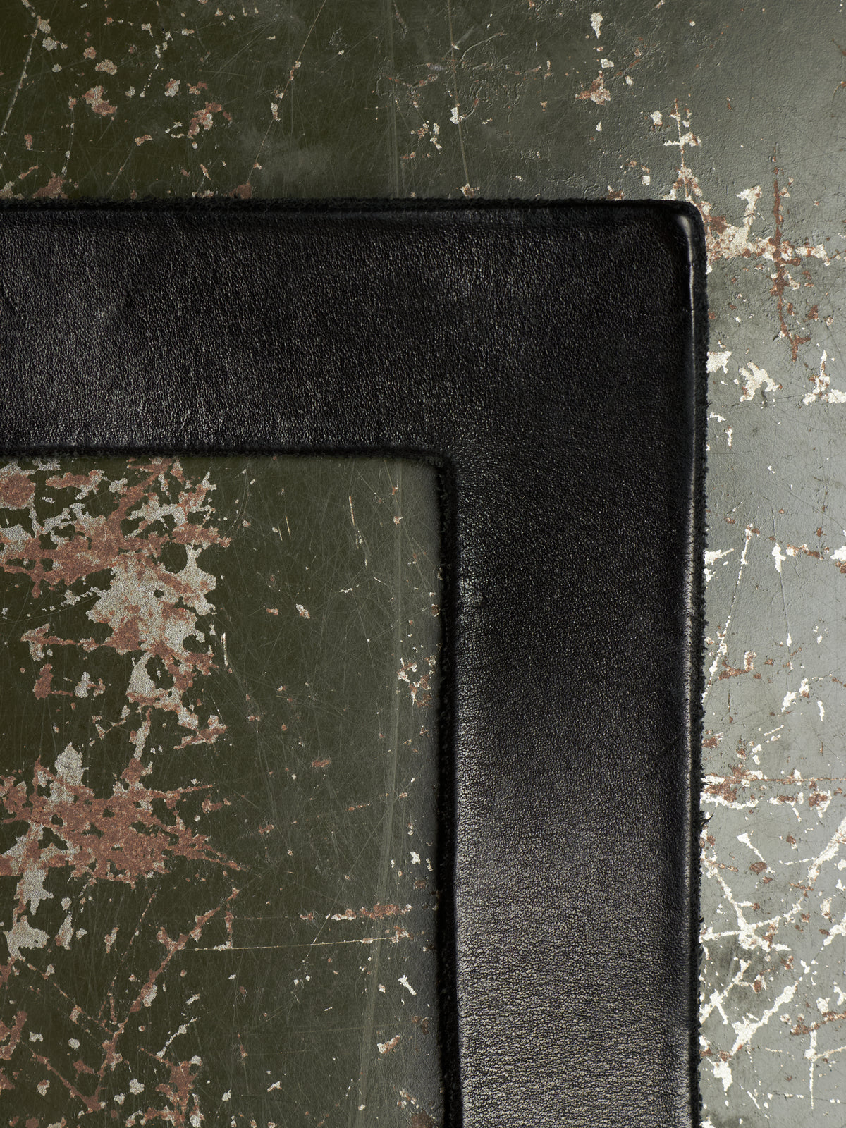 A close-up of a black leather or faux-leather rectangular frame corner resting on a distressed dark green surface with peeling white and brown paint.