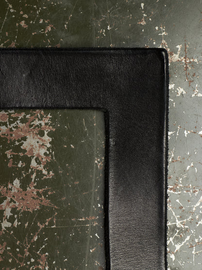 A close-up of a black leather or faux-leather rectangular frame corner resting on a distressed dark green surface with peeling white and brown paint.