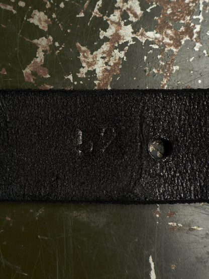 A close-up of a black textured leather belt strap laid horizontally across a distressed green and brown metal surface.