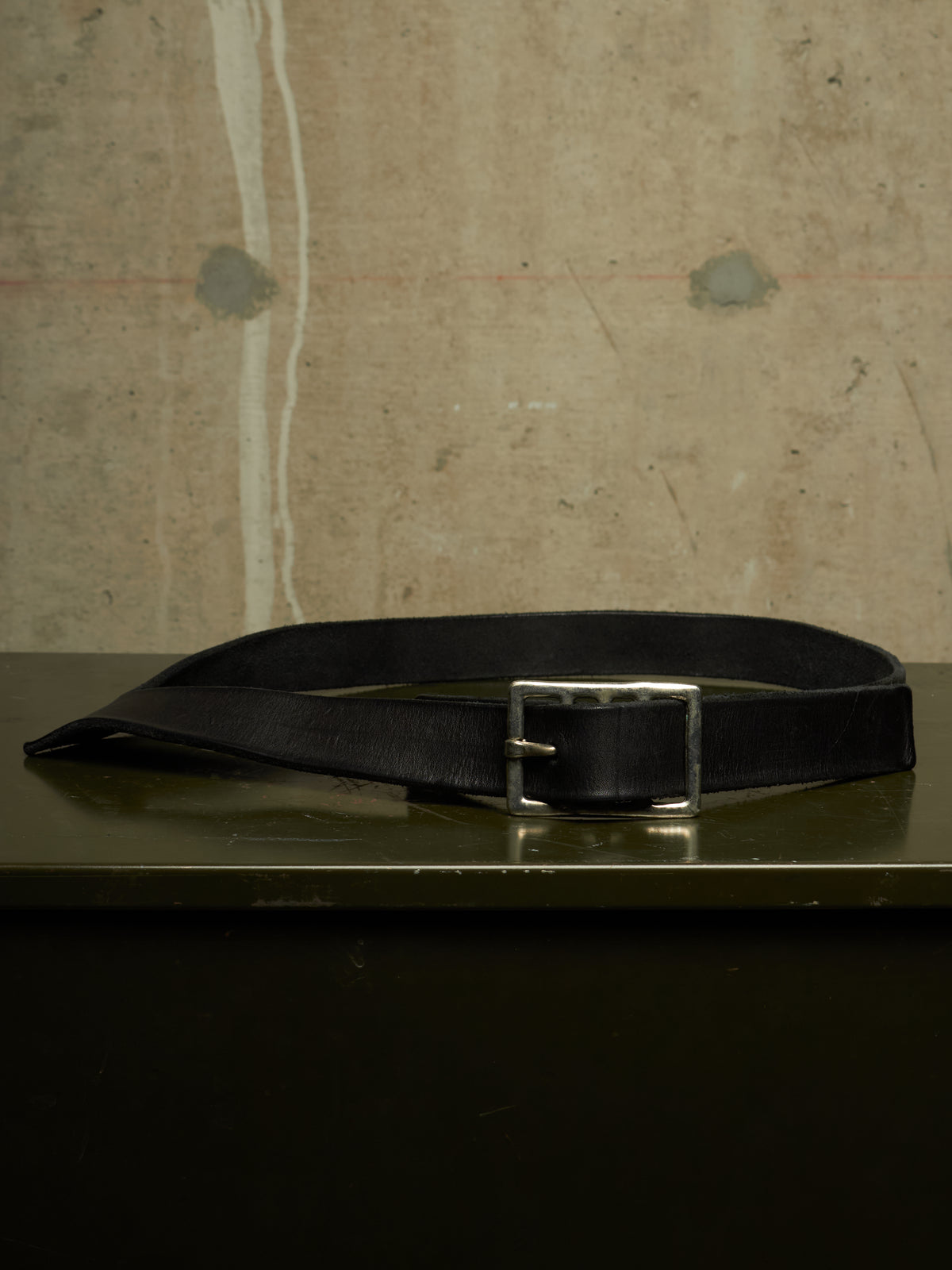 A black leather belt with a silver-toned metal buckle, resting on a dark reflective surface against a textured concrete wall background.