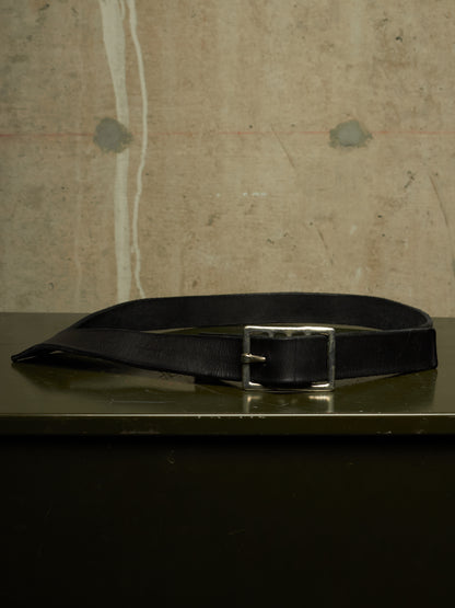 A black leather belt with a silver-toned metal buckle, resting on a dark reflective surface against a textured concrete wall background.