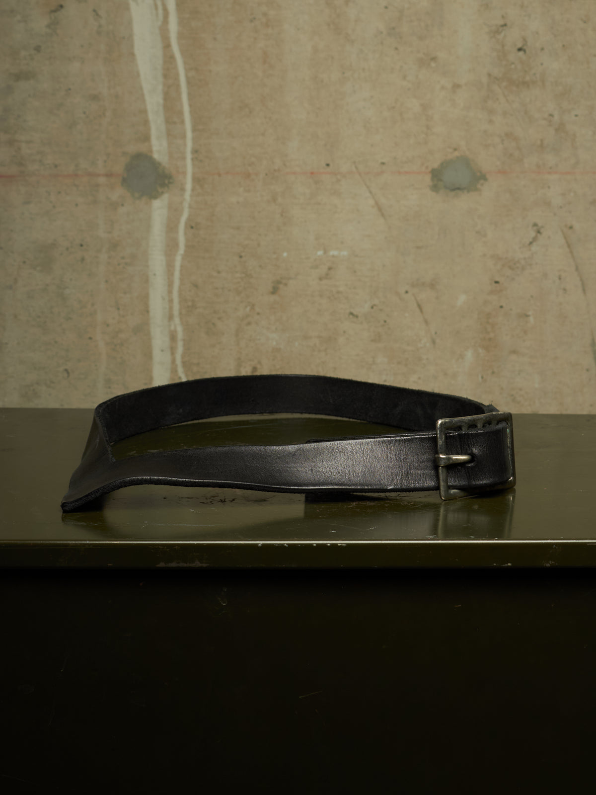 A black leather belt with a silver-toned metal buckle, resting on a dark reflective surface against a textured concrete wall background.