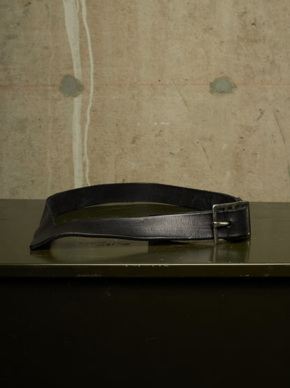 A black leather belt with a silver-toned metal buckle, resting on a dark reflective surface against a textured concrete wall background.