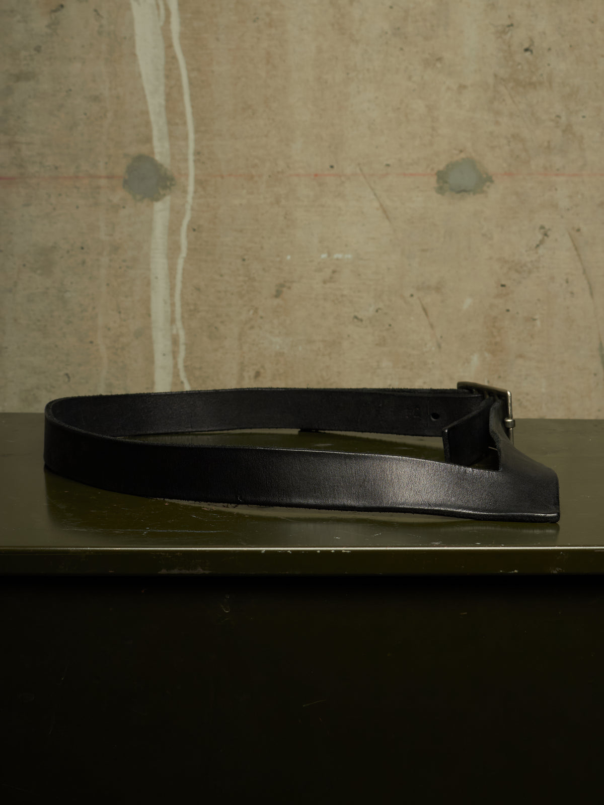 A black leather belt with a wide, structured strap and a dark metal buckle, displayed on a dark green surface against a textured concrete wall.