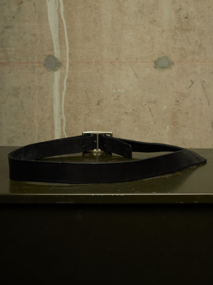A black leather belt with a silver-toned metal buckle, coiled on a dark green surface against a textured concrete wall background.