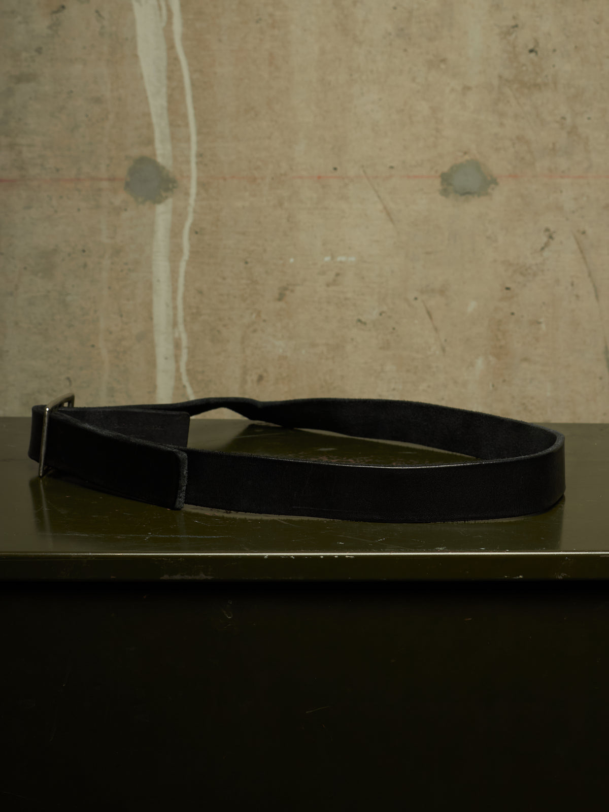 A black leather belt with a silver buckle, resting on a dark green surface against a textured concrete wall.